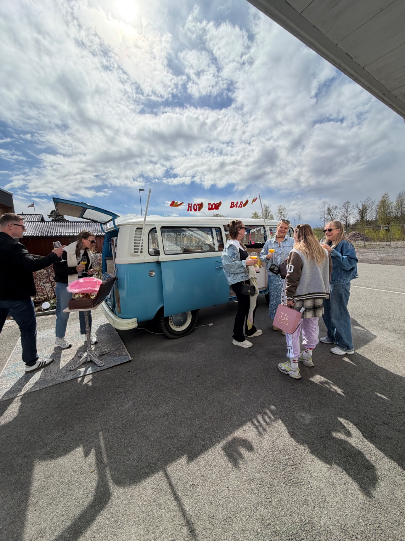 Gjester i denim og 90-tallsantrekk med drinker foran lyseblå VW-buss med Hot Dog Bar-banner