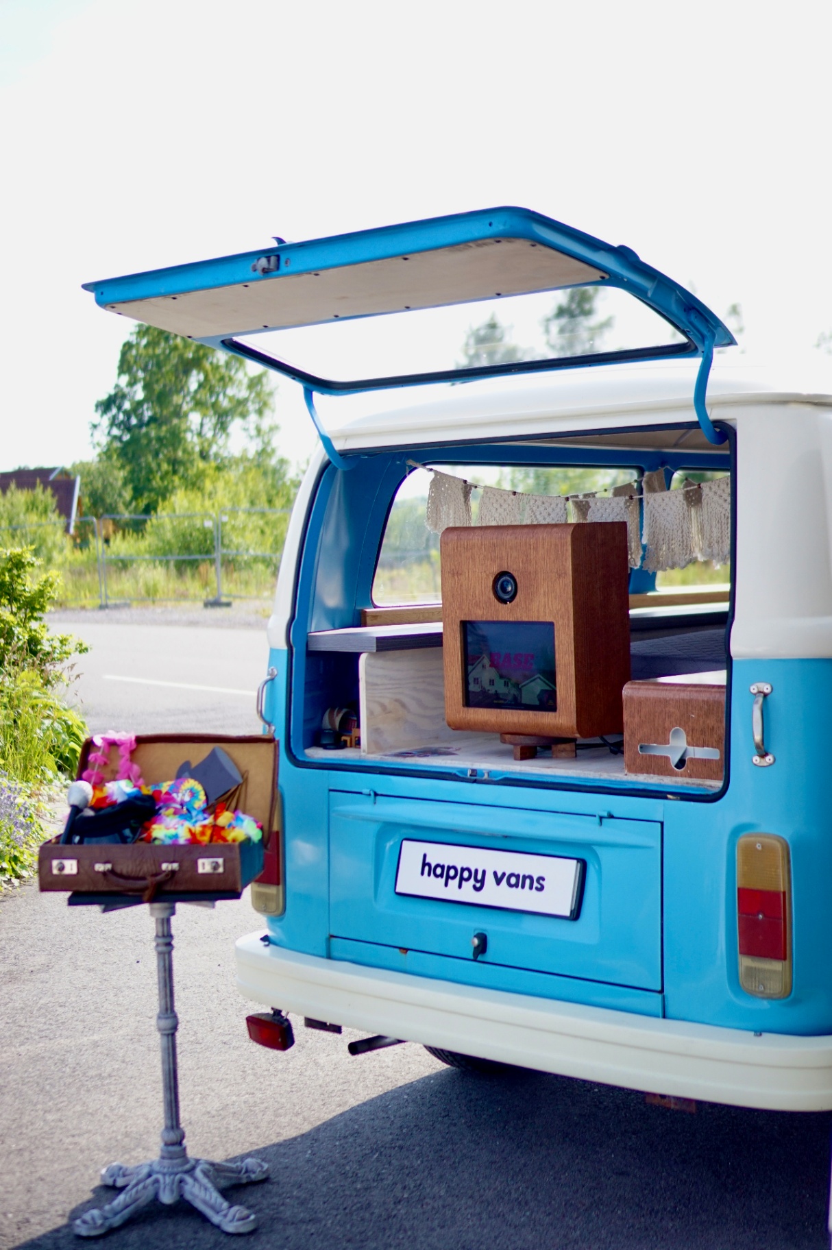 Lyseblå VW-buss med åpen baklukе, HappySmiles photobooth og en koffert med fargerike rekvisitter klar for gjestene